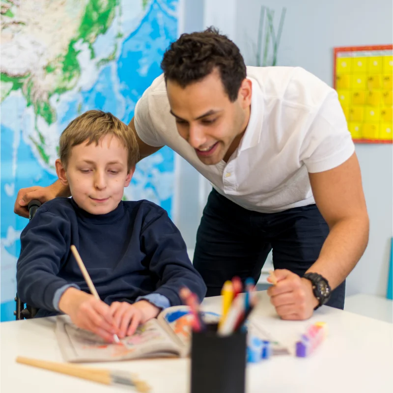 پسر بچه اتیستیک که با تمرکز در حال نوشتن است و مربی مرد کنارش آموزش می‌دهد