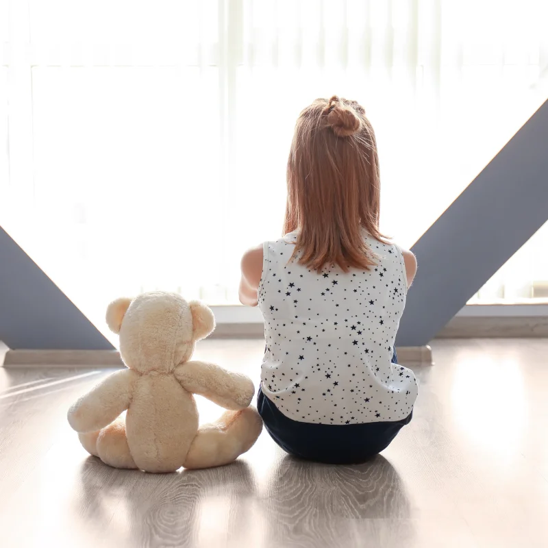 دختری با عروسکش پشت به دوربین نشسته و از پنجره بیرون را نگاه می‌کند، نمادی از تنهایی و دنیای درونی کودک