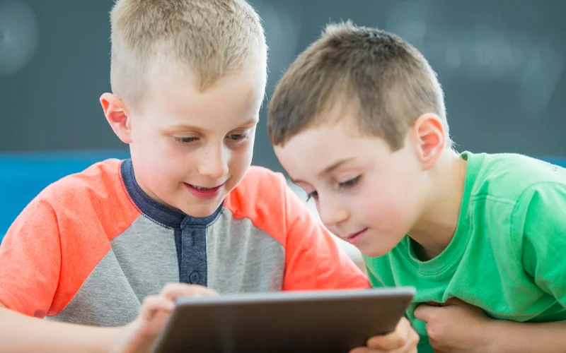  یکی از پسرها در حال بازی با تبلت و دیگری در حال تماشای صفحه برای یادگیری از بازی آموزشی