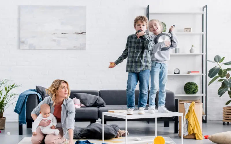 دو کودک بیش‌فعال که روی مبل ایستاده‌اند و با صدای بلند آواز می‌خوانند در حالی که مادر با نوزاد در آغوش به آن‌ها نگاه می‌کند