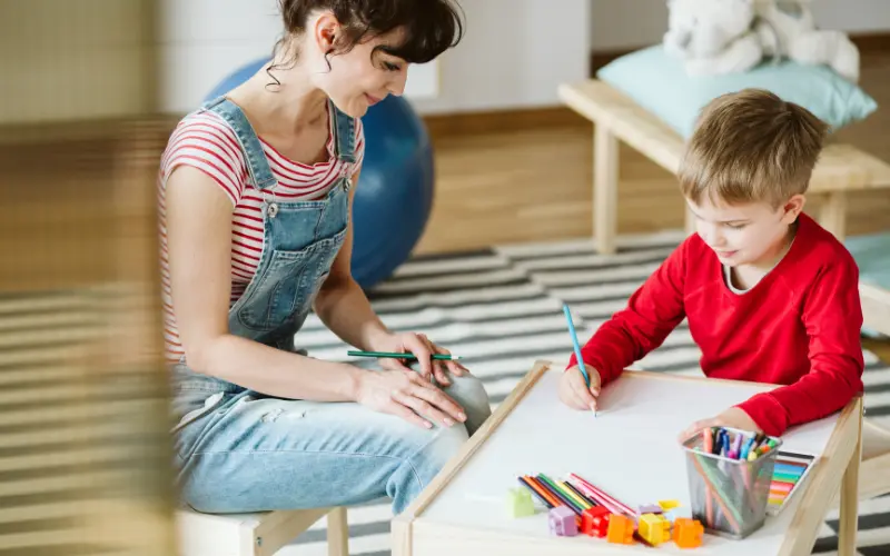 مادری که آرام و با تمرکز کنار کودک بیش‌فعالش نشسته و کودک در حال نقاشی کردن است